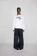 A model stands against a light gray studio background wearing a white long-sleeve sweatshirt with a black graphic print on the chest, paired with loose black pants and black sneakers.