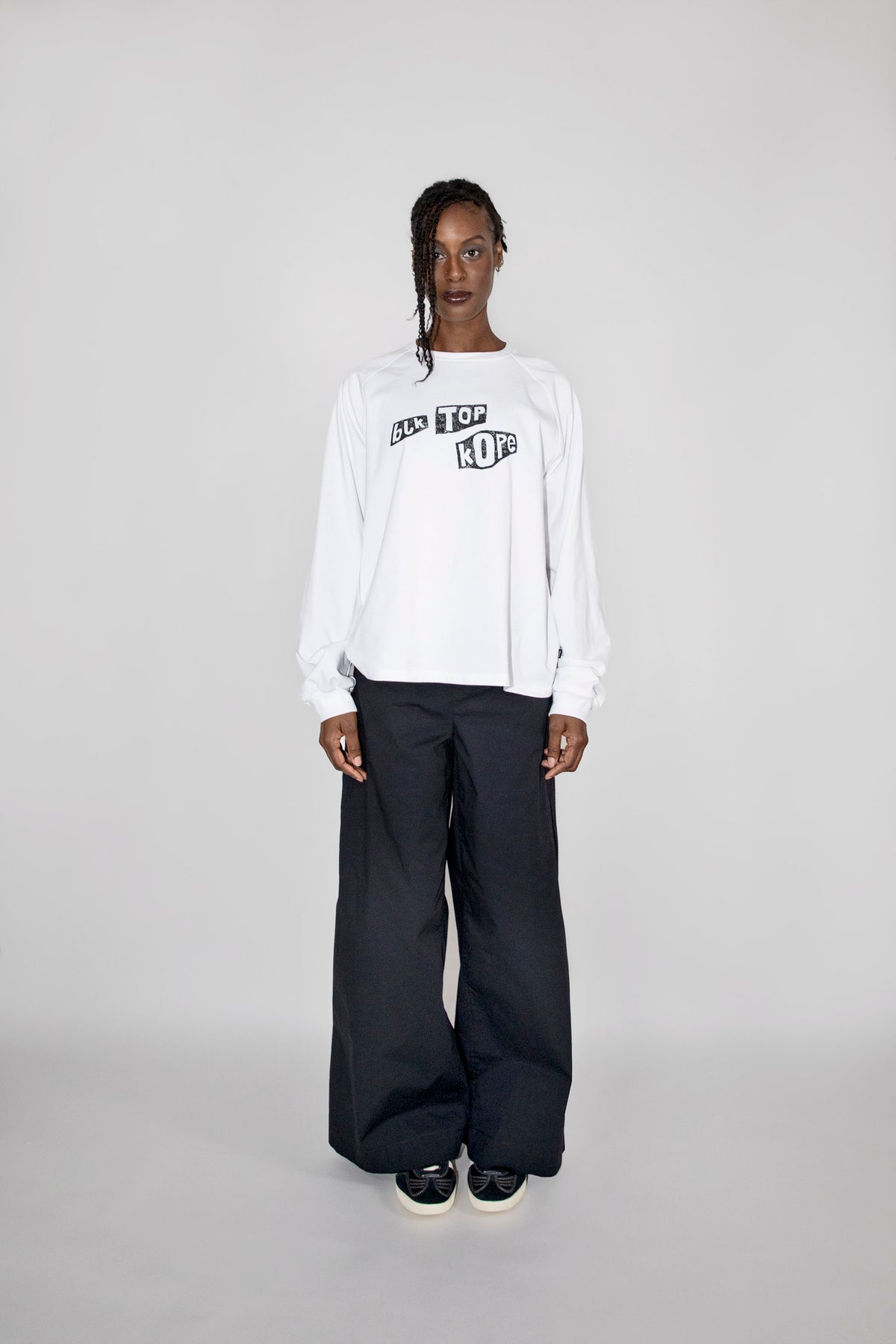 A model stands against a light gray studio background wearing a white long-sleeve sweatshirt with a black graphic print on the chest, paired with loose black pants and black sneakers.