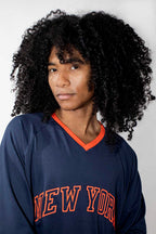 A person with voluminous curly black hair wearing a navy blue mesh long-sleeve jersey with orange trim and 'NEW YORK' embroidered across the chest, photographed against a light gray studio background.