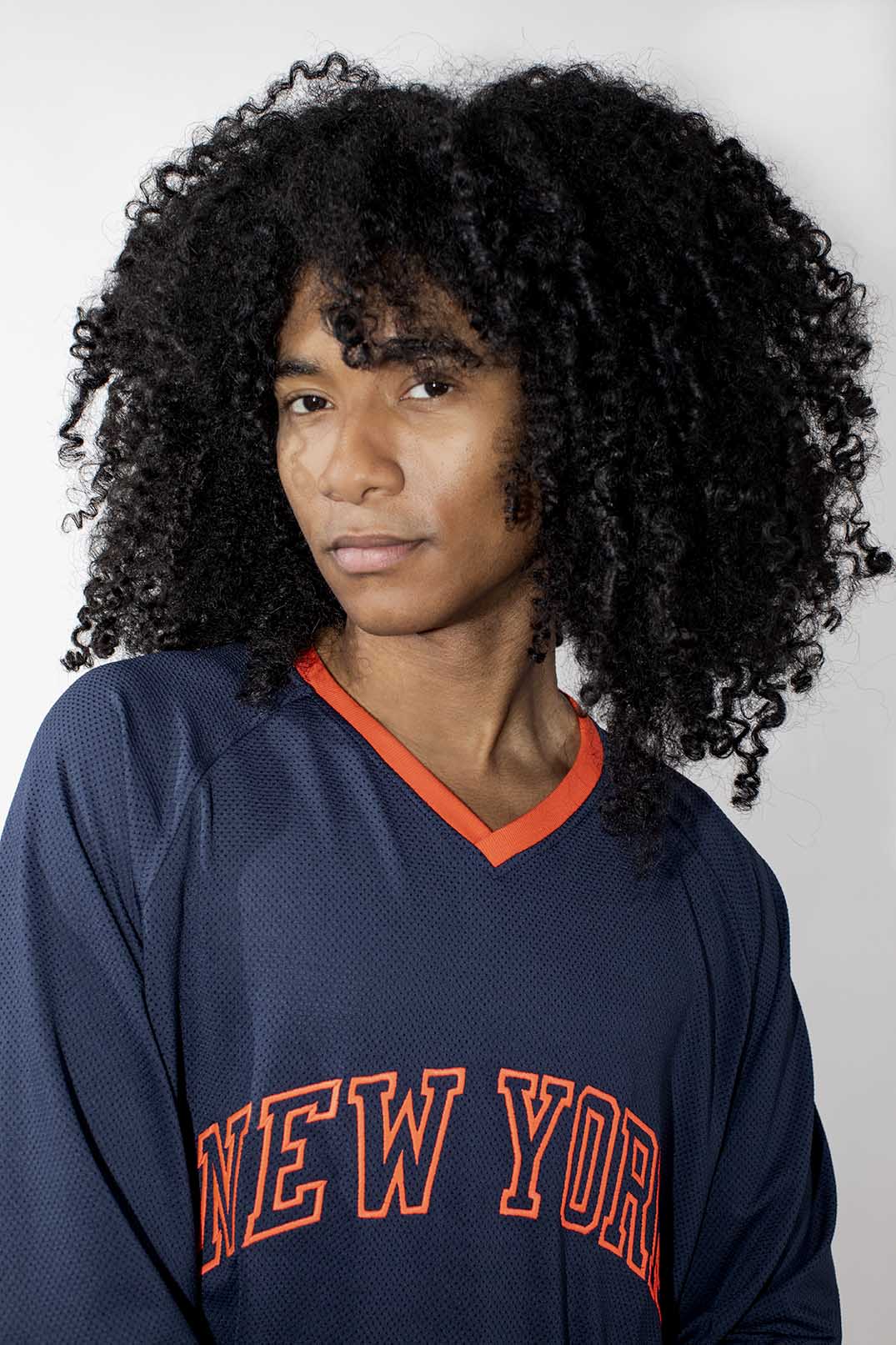 A person with voluminous curly black hair wearing a navy blue mesh long-sleeve jersey with orange trim and 'NEW YORK' embroidered across the chest, photographed against a light gray studio background.