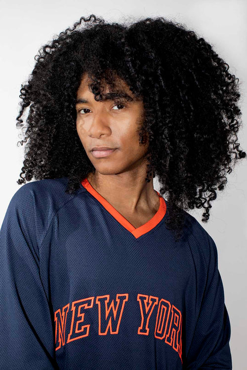 A person with voluminous curly black hair wearing a navy blue mesh long-sleeve jersey with orange trim and 'NEW YORK' embroidered across the chest, photographed against a light gray studio background.