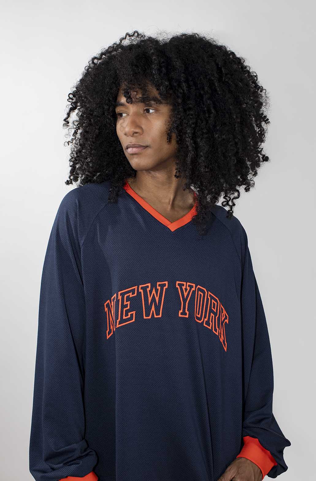 A person with curly black hair wearing an oversized navy blue long-sleeve 'NEW YORK' jersey-style top with orange trim and orange lettering, photographed against a light gray studio background.