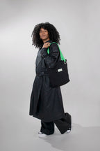 A model with curly black hair is shown from the side, wearing a long black trench coat, wide-leg black pants, and white sneakers, while carrying a black tote bag with a bright green strap against a light gray studio background.