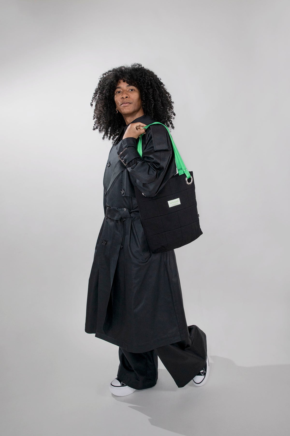 A model with curly black hair is shown from the side, wearing a long black trench coat, wide-leg black pants, and white sneakers, while carrying a black tote bag with a bright green strap against a light gray studio background.