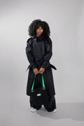 A model with curly black hair stands in a studio wearing a long black trench coat, wide-leg black pants, and white sneakers, while holding a black tote bag with bright green handles.