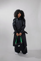 A model with curly black hair stands in a studio wearing a long black trench coat, wide-leg black pants, and white sneakers, while holding a black tote bag with bright green handles.