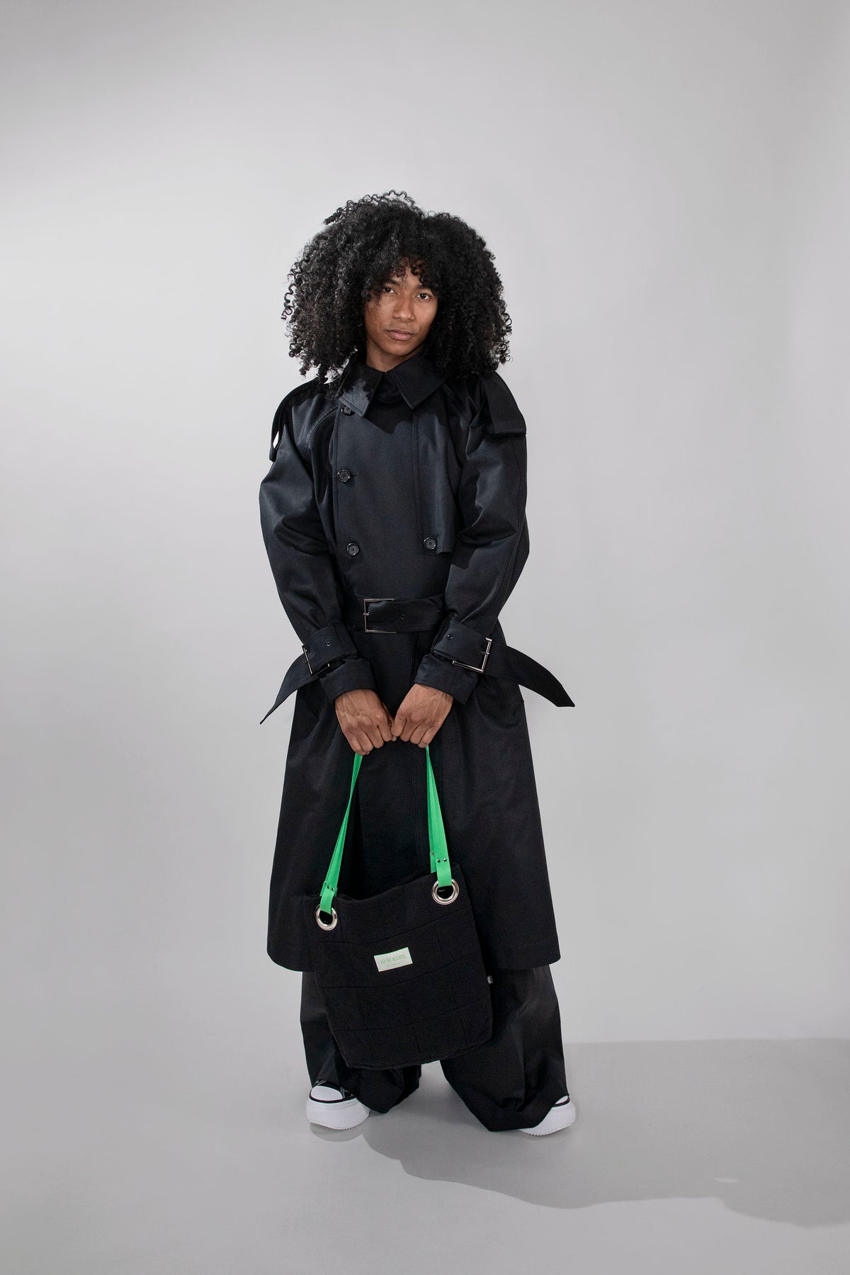 A model with curly black hair stands in a studio wearing a long black trench coat, wide-leg black pants, and white sneakers, while holding a black tote bag with bright green handles.