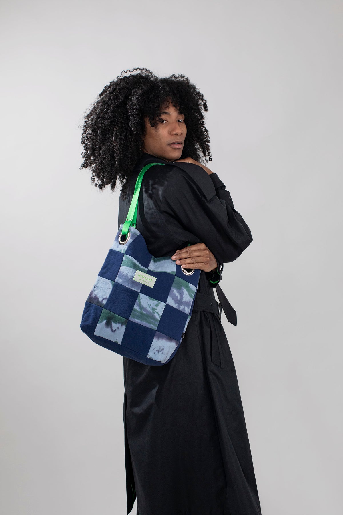 A person with curly black hair wearing a black trenchcoat, carrying a blue and tie dye patchwork canvas tote bag with a bright green strap, posed against a light gray studio background.