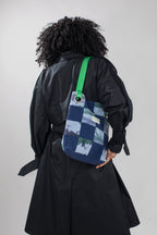 A person viewed from behind wearing a black trench coat and carrying a blue patchwork shoulder bag with a bright green strap against a light gray studio background.