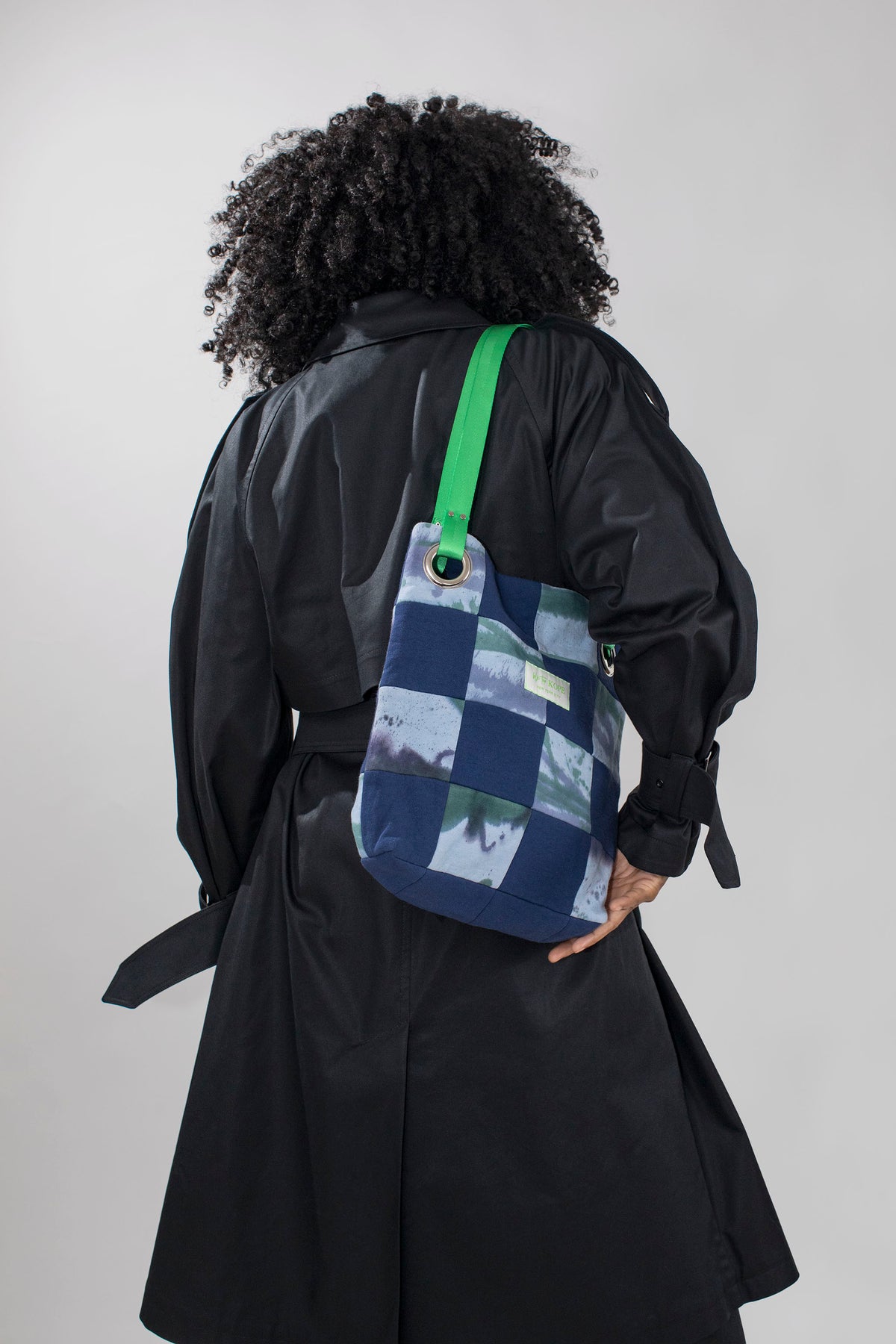 A person viewed from behind wearing a black trench coat and carrying a blue patchwork shoulder bag with a bright green strap against a light gray studio background.