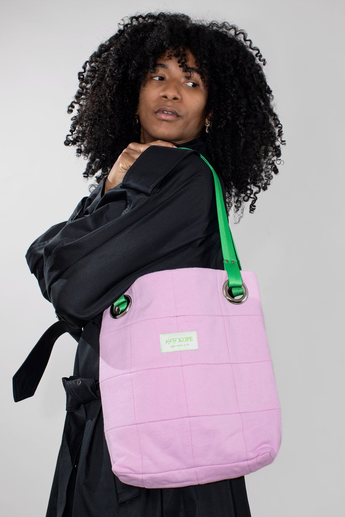 A person with curly black hair wearing a black coat carries a blush patchwork tote bag with a bright green shoulder strap and a small white brand label.