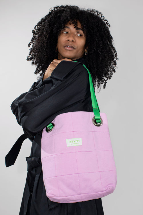A person with curly black hair wearing a black coat carries a blush patchwork tote bag with a bright green shoulder strap and a small white brand label.