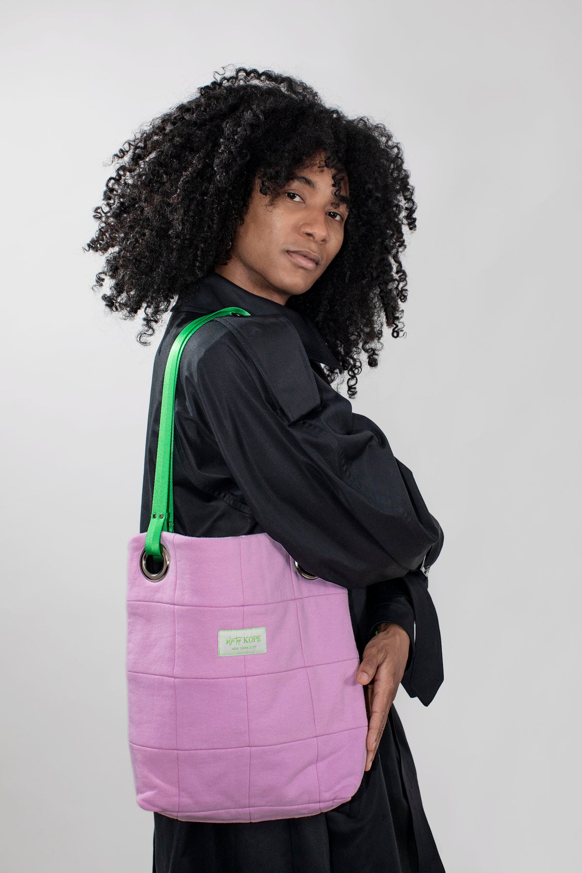 A model with curly black hair wearing a black trenchcoat and holding a blush patchwork tote bag with a bright green shoulder strap against a light gray background.