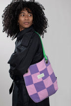A model with curly black hair wearing a black jacket, carrying a purple and pink checkered tote bag with a green strap against a light gray background.
