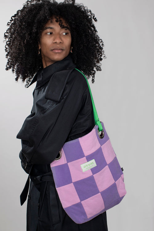 A model with curly black hair wearing a black jacket, carrying a purple and pink checkered tote bag with a green strap against a light gray background.