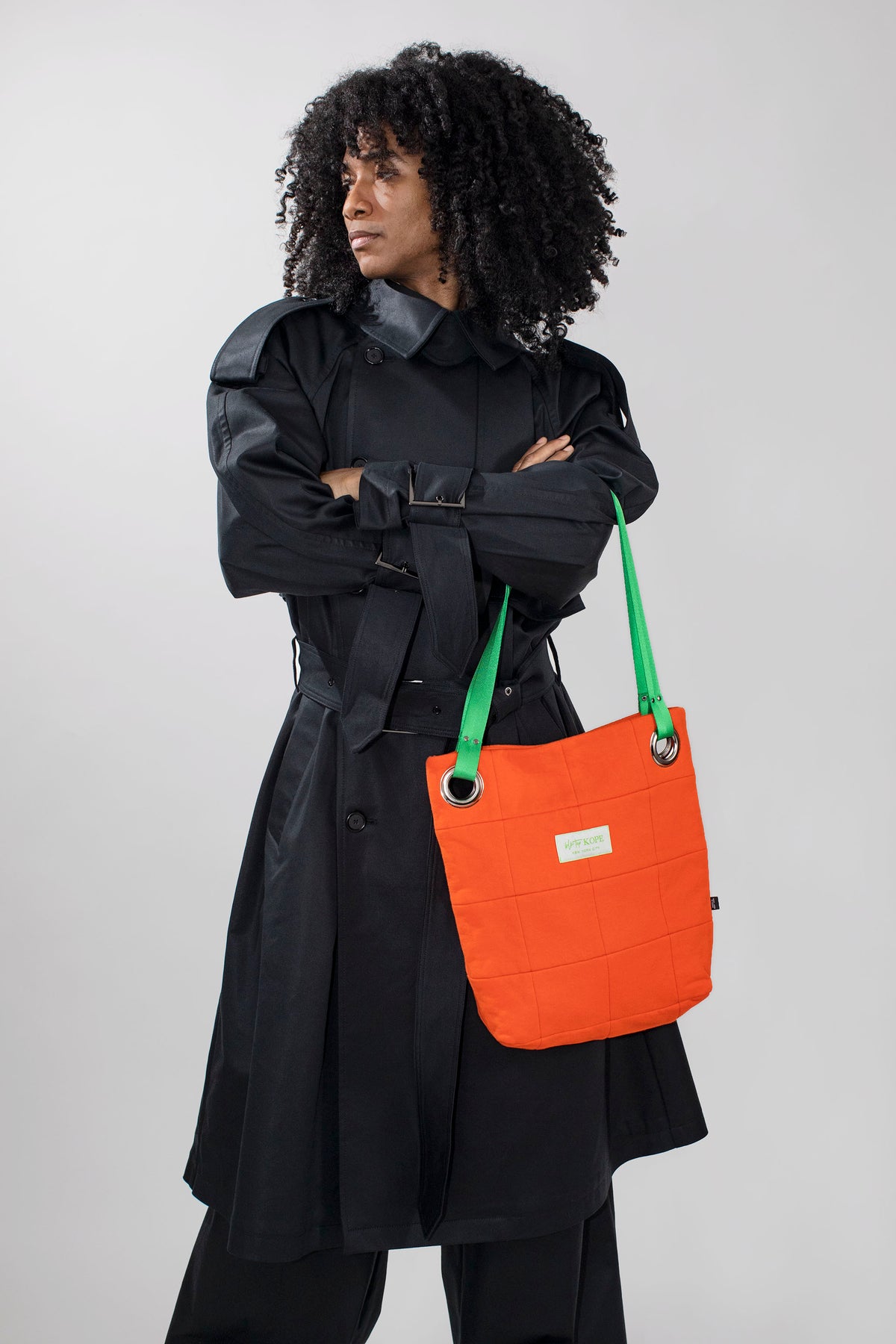 A model with curly black hair wearing a black trench coat and holding a bright orange canvas tote bag with green shoulder straps against a light gray studio background.