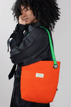 A model wearing a black jacket carries a bright orange quilted tote bag with a lime green shoulder strap and a small white label.