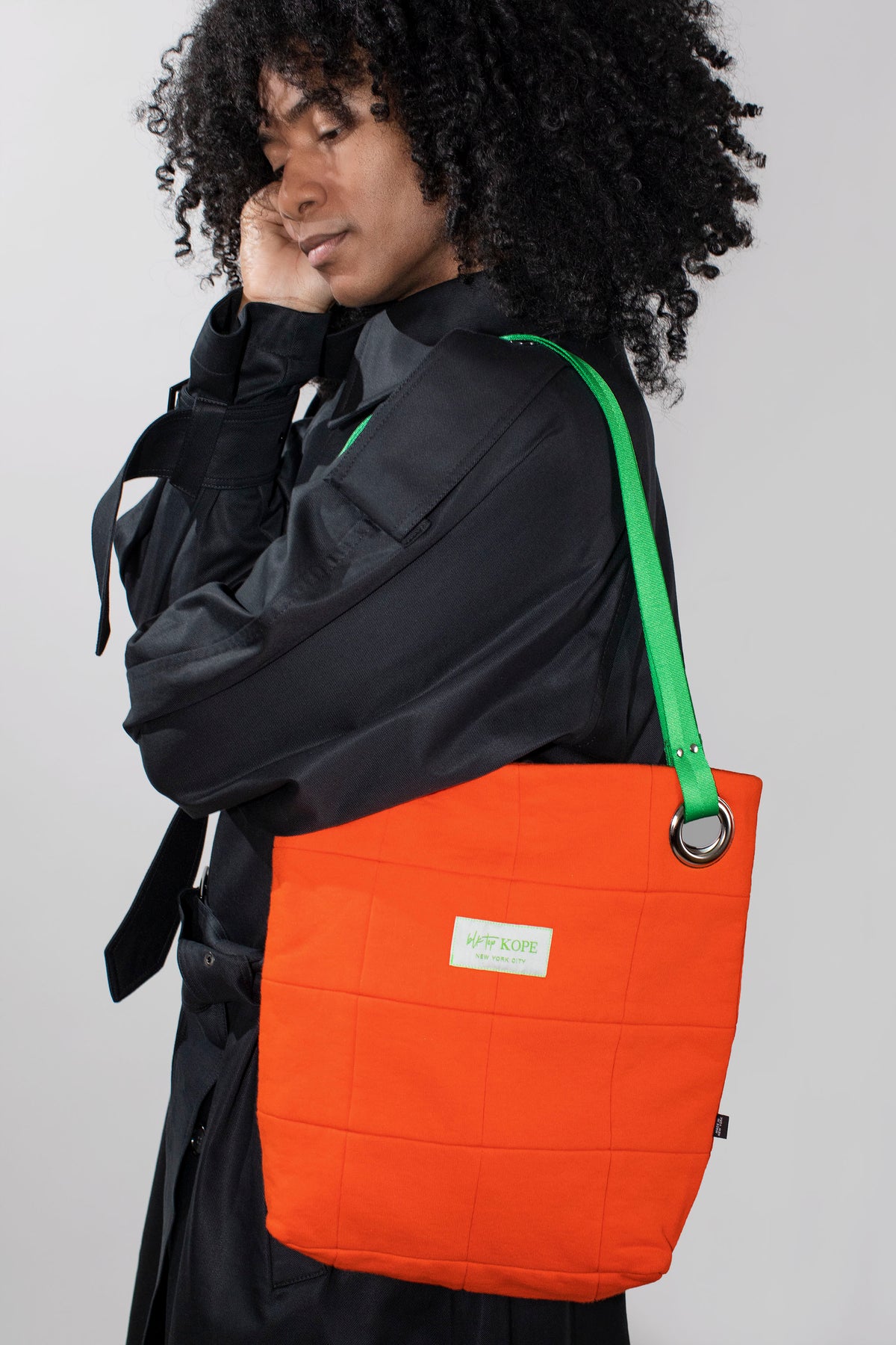 A model wearing a black jacket carries a bright orange quilted tote bag with a lime green shoulder strap and a small white label.