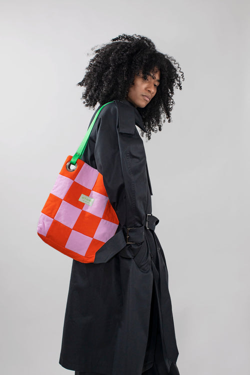 A model with curly black hair wearing a black trench coat, carrying a large checkered tote bag in orange and pink with a green strap, against a light gray background.