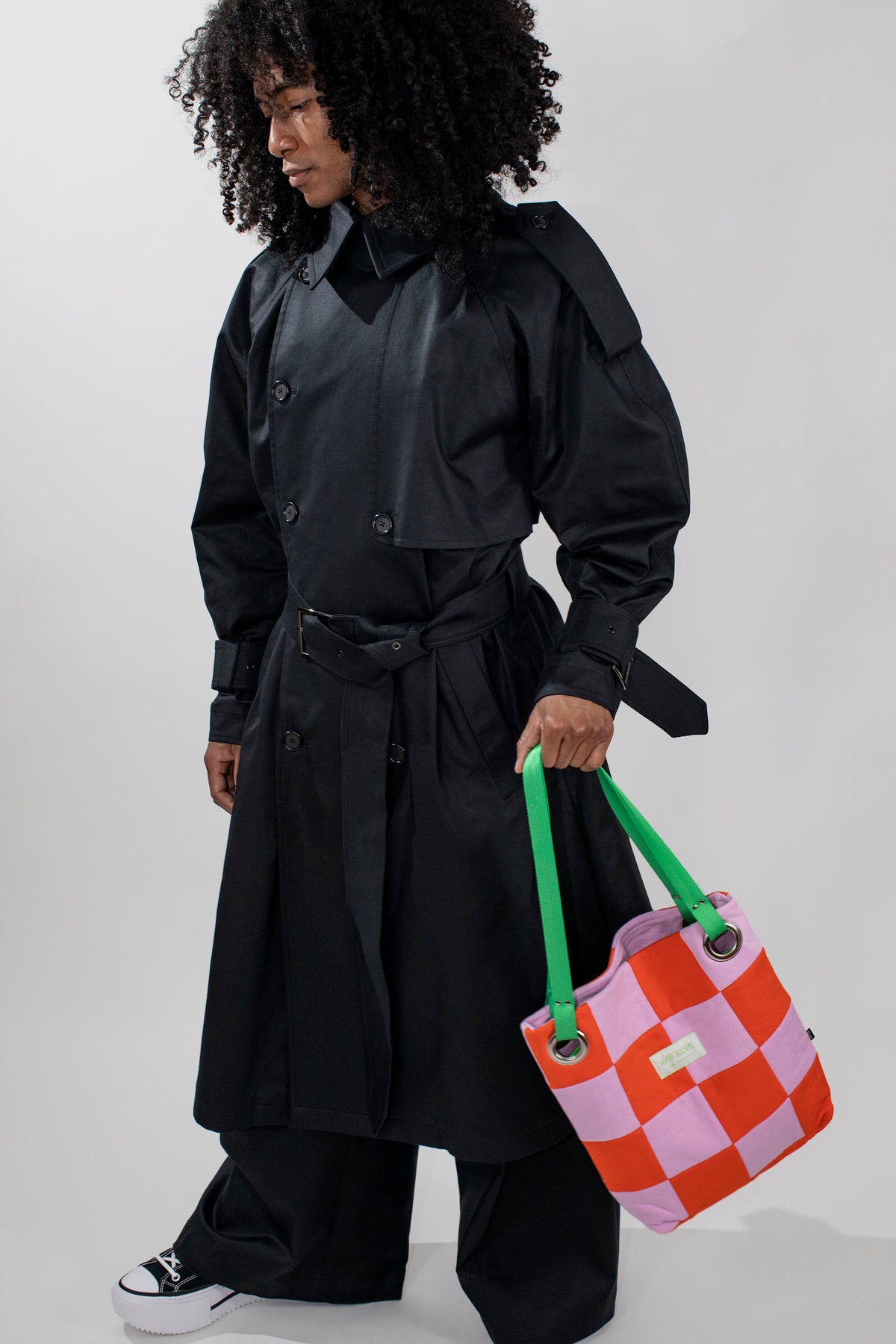 A model with curly black hair wearing a black oversized longline coat, black pants, and black sneakers, holding a pink and orange checkered tote bag with green handles against a light gray background.
