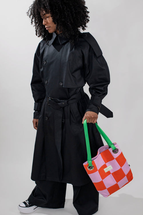 A model with curly black hair wearing a black oversized longline coat, black pants, and black sneakers, holding a pink and orange checkered tote bag with green handles against a light gray background.