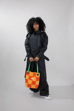 A model with curly black hair stands against a light gray studio background, wearing a black double-breasted trench coat and holding a patchwork orange and yellow checkered tote bag with green handles.