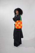 A model with curly black hair stands in profile wearing a black outfit and carrying a colorful checkered shoulder bag. The bag features an orange, and yellow checkerboard pattern with a green strap, set against a plain light gray studio background.