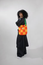 A model with curly black hair stands in profile wearing a black outfit and carrying a colorful checkered shoulder bag. The bag features an orange, and yellow checkerboard pattern with a green strap, set against a plain light gray studio background.