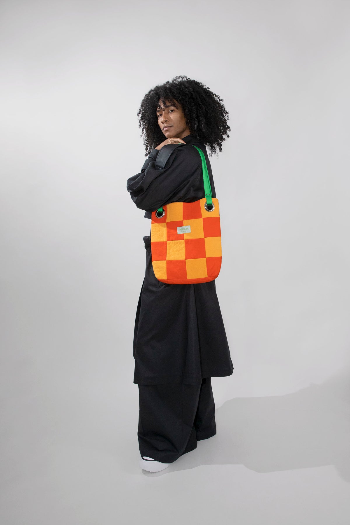 A model with curly black hair stands in profile wearing a black outfit and carrying a colorful checkered shoulder bag. The bag features an orange, and yellow checkerboard pattern with a green strap, set against a plain light gray studio background.