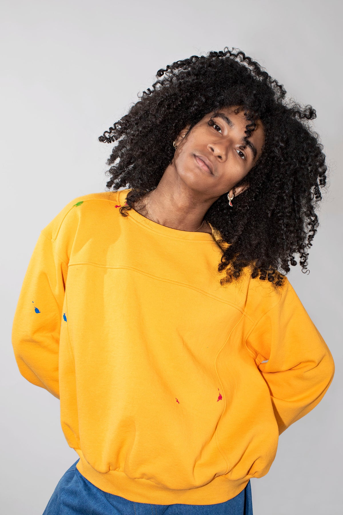 A person with voluminous curly black hair wearing a yellow oversized sweatshirt with small colorful paint splatter details, paired with blue jeans, photographed against a plain light gray background.