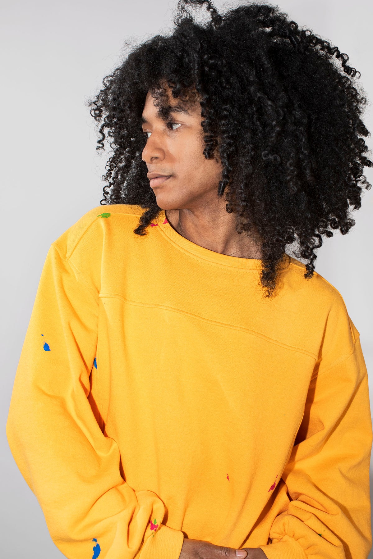 A person with voluminous black curly hair wearing a yellow sweatshirt with small multicolor paint splatters, photographed against a plain light gray studio background.