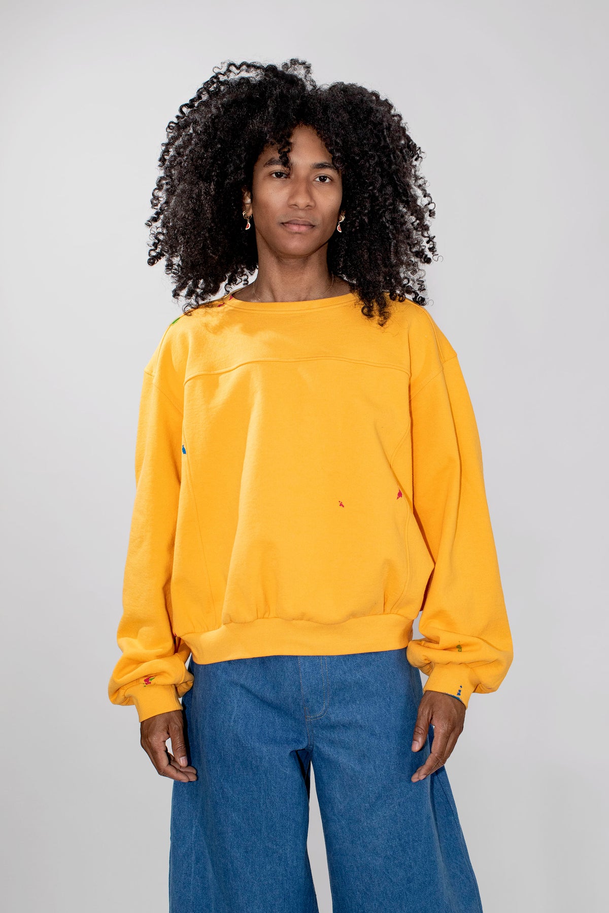 A model with curly black hair wearing a yellow oversized crewneck sweatshirt and blue wide-leg jeans, standing against a light gray studio background.
