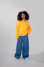 A model with curly black hair stands against a light gray studio background wearing a yellow oversized sweatshirt, wide-leg blue jeans, and white sneakers.