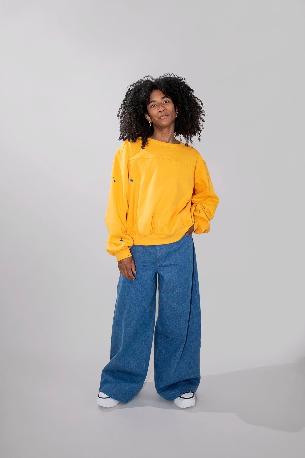 A model with curly black hair stands against a light gray studio background wearing a yellow oversized sweatshirt, wide-leg blue jeans, and white sneakers.
