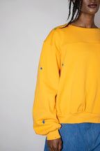 A close-up of a person wearing a yellow crewneck sweatshirt with multicolored paint splatters on the chest and sleeves, paired with blue jeans against a plain light gray background.