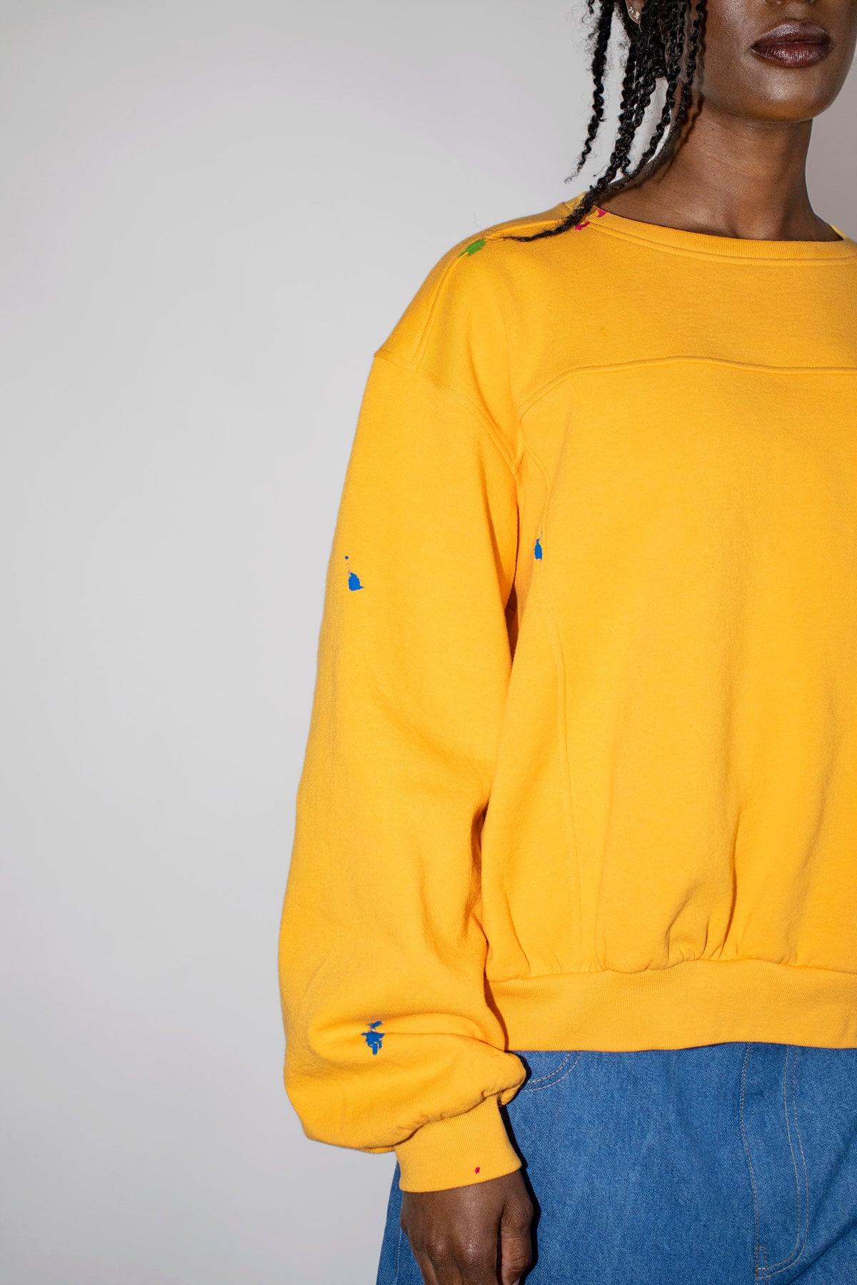 A close-up of a person wearing a yellow crewneck sweatshirt with multicolored paint splatters on the chest and sleeves, paired with blue jeans against a plain light gray background.