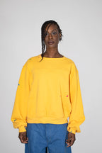 A model wearing a yellow oversized crewneck sweatshirt with small colorful paint splatter details on the sleeves and front, paired with blue jeans, against a plain light gray background.