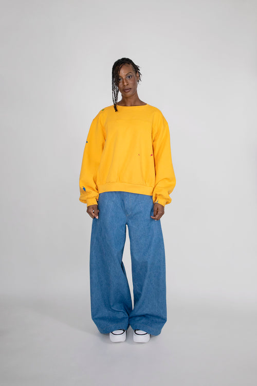 A model stands against a light gray studio background wearing a yellow crewneck sweatshirt and very wide-leg blue jeans with white sneakers.