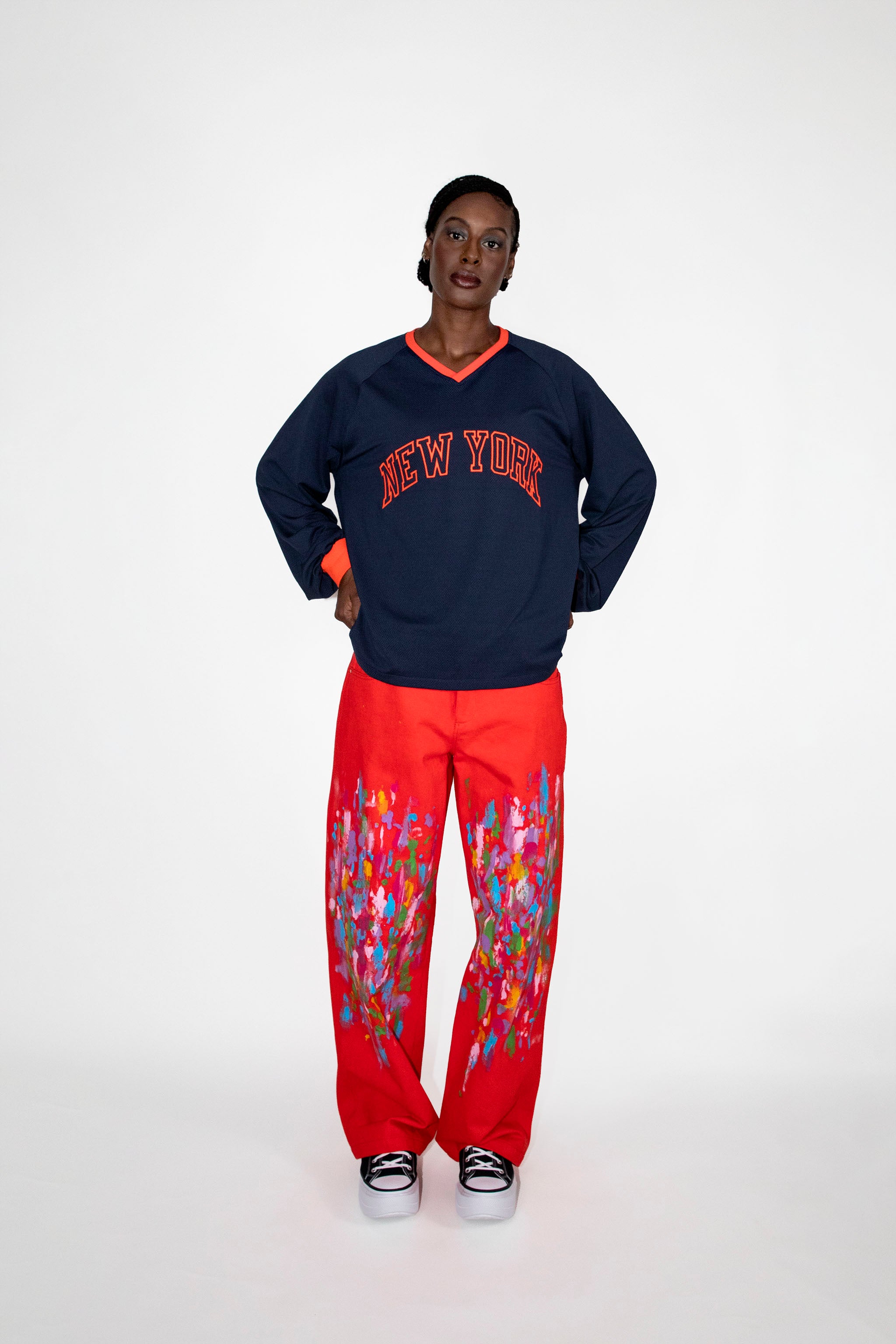 Person wearing a navy sweatshirt with 'New York' text and red pants with colorful paint on a white background