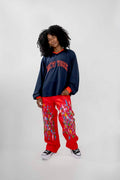 A model with curly black hair stands against a light gray studio background wearing a navy blue long-sleeve crewneck sweatshirt with 'NEW YORK' embroidered in red across the chest, paired with bright red cargo-style pants featuring a colorful paint pattern, and black-and-white sneakers.