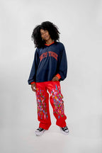 A model with curly black hair stands against a light gray studio background wearing a navy blue long-sleeve crewneck sweatshirt with 'NEW YORK' embroidered in red across the chest, paired with bright red cargo-style pants featuring a colorful paint pattern, and black-and-white sneakers.
