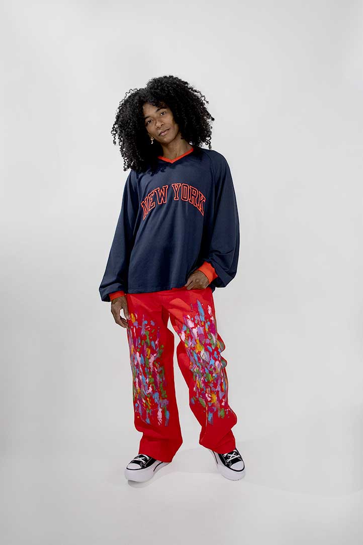 A model with curly black hair stands against a light gray studio background wearing a navy blue long-sleeve crewneck sweatshirt with 'NEW YORK' embroidered in red across the chest, paired with bright red cargo-style pants featuring a colorful paint pattern, and black-and-white sneakers.