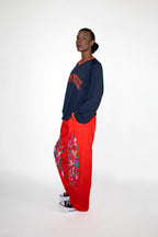 Person wearing a navy sweatshirt with red "New York" text and red pants with paint patterns on a white background