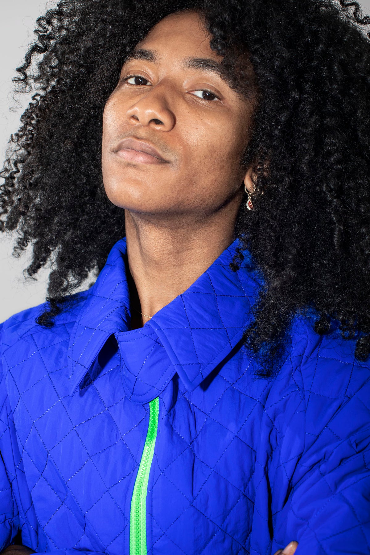 A close-up portrait of a person with voluminous dark curly hair wearing a bright cobalt blue quilted jacket with a neon green zipper, set against a plain light gray background.