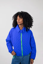 A model with curly black hair wearing a cobalt blue quilted jacket with a neon green zipper, paired with blue jeans, against a plain light gray background.