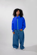 A model with curly black hair stands against a light gray studio background wearing a cobalt blue quilted jacket with a neon green zipper, paired with wide-leg blue jeans and white sneakers.