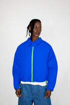 A model wearing a cobalt blue quilted jacket with a neon green front zipper, paired with blue jeans, against a plain light gray background.