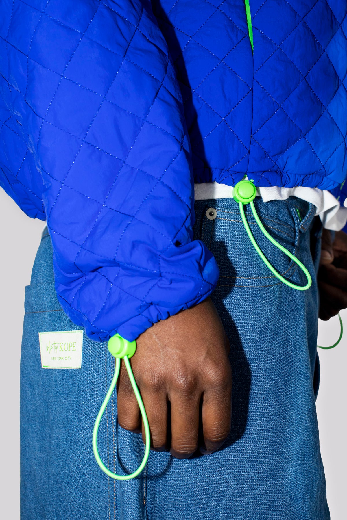 Close-up of a person wearing a cobalt blue quilted jacket with neon green drawstring loops and blue jeans, showing the sleeve cuff and hand area.