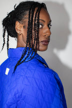 A woman with black braided hair wearing a cobalt blue quilted jacket, shown in a close-up side profile against a plain light gray wall.