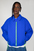 A model wearing a bright cobalt blue quilted jacket with a neon green zipper and matching drawstrings, paired with a white undershirt and blue jeans, against a plain light gray background.